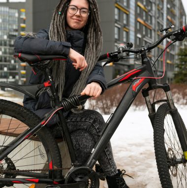 Photo on the street, a young active woman with a dreadlocked hairstyle, dressed in a warm jacket, rides a bicycle.